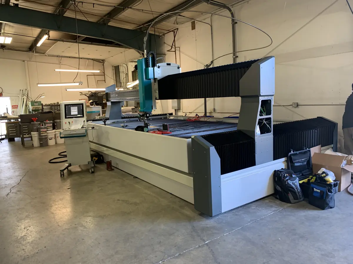 A Flow CNC waterjet cutting system on a shop floor during installation, gantry and bellows way covers fitted, control pendant alongside the slat table.