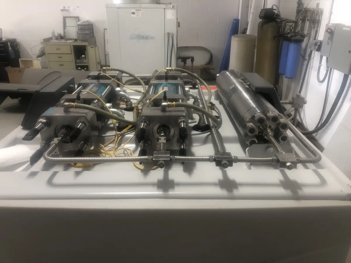 High-pressure intensifier assemblies and an accumulator laid out on a bench during a pump rebuild, water treatment equipment behind.